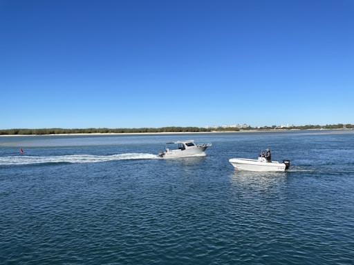 Two motorboats are gliding across a body of water under a clear blue sky. The scene includes one boat trailing white foamy water behind it, while the other is moving alongside. The horizon features a sandy shoreline with green shrubs and a distant view of buildings against the blue sky.