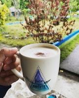 A hand holding a white mug with the words 'free spirit' printed on it. The mug contains a frothy beverage, possibly coffee, with a sprinkle of cocoa or cinnamon on top. The background features a vibrant outdoor setting with greenery and red foliage, suggesting a garden or yard.