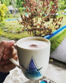 A hand holding a white mug with the words 'free spirit' printed on it. The mug contains a frothy beverage, possibly coffee, with a sprinkle of cocoa or cinnamon on top. The background features a vibrant outdoor setting with greenery and red foliage, suggesting a garden or yard.