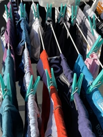 Various colorful clothes are hung to dry on a drying rack, secured with teal clothespins. The clothes are in shades of red, blue, black, white, and gray.
