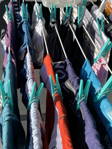 Various colorful clothes are hung to dry on a drying rack, secured with teal clothespins. The clothes are in shades of red, blue, black, white, and gray.