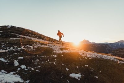 A person wearing a red jacket is running uphill on a rugged terrain covered partially with snow and rocks. The sun is setting or rising in the background, casting a warm golden light across the scene. The horizon displays a range of mountains under a clear sky.