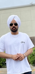 A person wearing a white turban, white T-shirt, and sunglasses stands outdoors. They have a beard and are wearing a necklace and a bracelet.