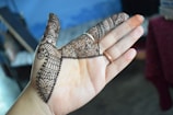 A hand with intricate henna designs covering the fingers and part of the palm. The patterns include swirls, lines, and dots, showing traditional artistic elements. The background is slightly blurred with a mix of blue and dark shades, suggesting an indoor environment.