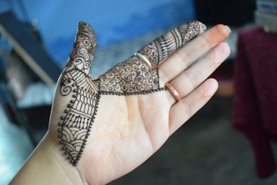 A hand with intricate henna designs covering the fingers and part of the palm. The patterns include swirls, lines, and dots, showing traditional artistic elements. The background is slightly blurred with a mix of blue and dark shades, suggesting an indoor environment.