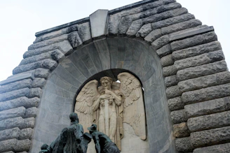 A stone relief sculpture depicting an angel with large wings holding a sword, set within a deeply recessed archway. Below the angel, three human figures carved from stone appear to be contemplating or approaching. The surrounding structure is composed of massive, rugged stone blocks arranged in an arch.