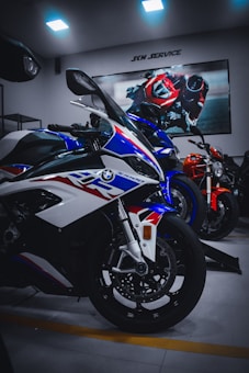 Several sports motorcycles are parked indoors, with a prominent white, blue, and red bike in the foreground. A poster of a racing motorcycle rider is displayed on the wall above them, and the room is lit by ceiling lights.