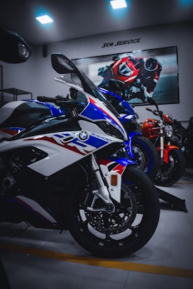 Several sports motorcycles are parked indoors, with a prominent white, blue, and red bike in the foreground. A poster of a racing motorcycle rider is displayed on the wall above them, and the room is lit by ceiling lights.