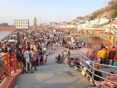 A large crowd gathers on a riverbank, likely for a cultural or religious event. The area is bustling with people of various ages in traditional and modern attire. In the background, there are orange footbridges and structures, along with a clock tower. The river flows to the left side of the image, and the sun casts a warm light over the scene.
