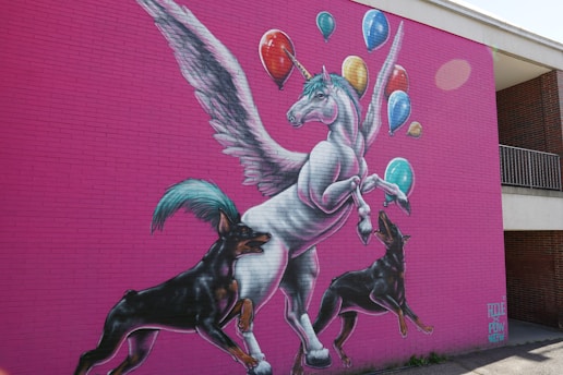 A vibrant mural on a pink brick wall depicts a winged unicorn with colorful balloons floating above. Two playful dogs with black fur are interacting with the unicorn, adding a sense of movement and whimsy.