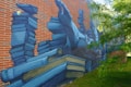 A mural painted on a brick wall depicting a large stack of blue-toned books. The artwork includes detailed illustrations of books in various positions, some open, creating a three-dimensional effect. There's a tree partially obscuring the right side of the mural.