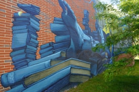A mural painted on a brick wall depicting a large stack of blue-toned books. The artwork includes detailed illustrations of books in various positions, some open, creating a three-dimensional effect. There's a tree partially obscuring the right side of the mural.