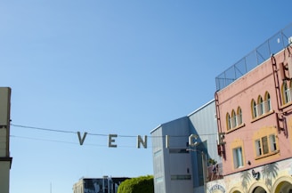 Metallic letters spelling out 'VENICE' are suspended across a street, with a clear blue sky in the background. To the right, there's a colorful building with arched windows and a mural, and to the left are modern structures.