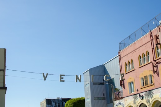 Metallic letters spelling out 'VENICE' are suspended across a street, with a clear blue sky in the background. To the right, there's a colorful building with arched windows and a mural, and to the left are modern structures.