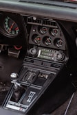The interior of a classic car featuring a detailed dashboard with multiple gauges, including RPM, water temperature, oil pressure, and battery level indicators. The audio system displays the Corvette logo, alongside volume and tuning controls. Below, the gear shift lever with a metallic knob is positioned next to a row of buttons for various vehicle functions.