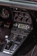 The interior of a classic car featuring a detailed dashboard with multiple gauges, including RPM, water temperature, oil pressure, and battery level indicators. The audio system displays the Corvette logo, alongside volume and tuning controls. Below, the gear shift lever with a metallic knob is positioned next to a row of buttons for various vehicle functions.