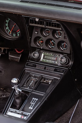 The interior of a classic car featuring a detailed dashboard with multiple gauges, including RPM, water temperature, oil pressure, and battery level indicators. The audio system displays the Corvette logo, alongside volume and tuning controls. Below, the gear shift lever with a metallic knob is positioned next to a row of buttons for various vehicle functions.