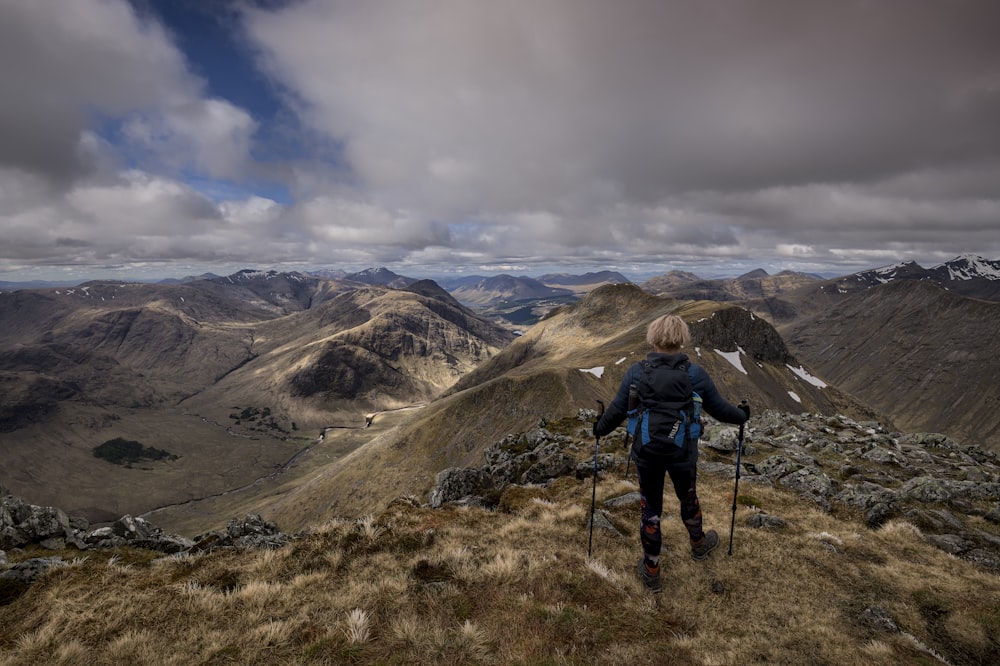 person in blue jacket and black pants with black hiking bag on mountain under white clouds