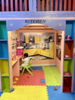 A child is seated on a small green chair at a colorful play kitchen setup. The kitchen features vivid colors with yellow cabinets depicted on the wall, a play stove, and various kitchen toys. The floor is covered with bright interlocking foam mats in blue, yellow, and red.
