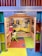 A child is seated on a small green chair at a colorful play kitchen setup. The kitchen features vivid colors with yellow cabinets depicted on the wall, a play stove, and various kitchen toys. The floor is covered with bright interlocking foam mats in blue, yellow, and red.
