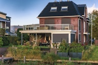 A modern two-story brick house with a dark sloped roof, featuring a large balcony with glass railings. The house is surrounded by a well-maintained garden with various plants and trees. There are three dogs siting on the lawn in front of the house, and a small pond or water feature is visible at the edge of the property. The scene appears to be set in a suburban neighborhood with other homes nearby.