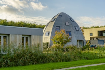 A modern, uniquely shaped dome house with multiple small windows is surrounded by lush greenery, including a tree with autumn-colored leaves. Adjacent to it, there are rectangular, modern structures with a sleek, minimalistic design. The sky is partially cloudy with soft, warm sunlight enhancing the scene.