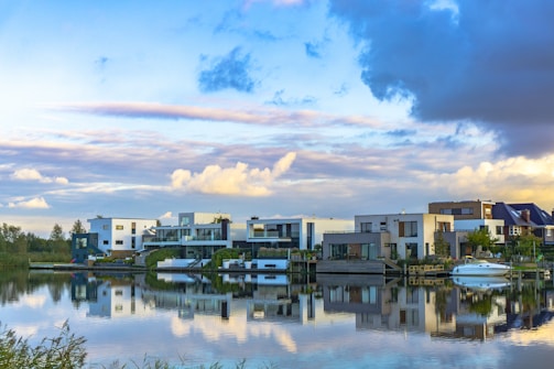 Modern waterfront houses are reflected in a calm body of water, with a partly cloudy sky above. The buildings are contemporary in style with large glass windows, and lush greenery surrounds them. A boat is docked at a pier adjacent to the homes, and the vibrant sky includes hues of blue, yellow, and gray.
