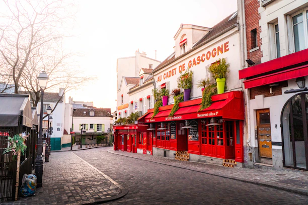 A European street scene featuring a charming restaurant with vibrant red accents named 'AU CADET DE GASCOGNE'. The restaurant's exterior is adorned with colorful potted plants. The street is cobblestone, and the buildings exhibit classic architectural styles with a mix of red, beige, and white tones. There are street lamps and minimal vegetation visible in the background.