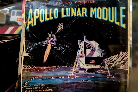 An illustration depicts the Apollo Lunar Module on the moon's surface, with stars visible in the background. The image includes the text 'APOLLO LUNAR MODULE' at the top and 'BATTERY OPERATED' above it. The Lunar Module displays the United States flag, and the backdrop features lunar landscapes and mountains.