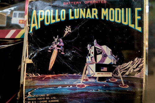 An illustration depicts the Apollo Lunar Module on the moon's surface, with stars visible in the background. The image includes the text 'APOLLO LUNAR MODULE' at the top and 'BATTERY OPERATED' above it. The Lunar Module displays the United States flag, and the backdrop features lunar landscapes and mountains.