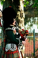 A person is wearing a traditional Scottish outfit, including a tartan kilt and a black fur hat, while playing the bagpipes. They stand outdoors next to a tree with a blurred background of foliage and a fence.