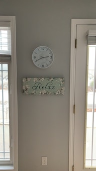 A minimalist interior wall featuring a round white clock showing the time as 9:10. Below the clock, there is a decorative rectangular sign with floral designs and the word 'Relax' written in cursive. A power outlet is visible near the bottom. The wall is painted a light grey color, and there is natural light coming through the adjacent window and glass door.