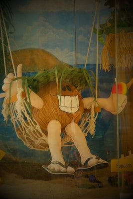 A whimsical coconut character with a large toothy smile is lounging on a hammock. It has arms and legs, wears flip-flops, and is topped with a leafy hat. The character is holding a colorful snow cone in one hand, surrounded by a beach-themed backdrop including the ocean, sand, and hills under a partly cloudy sky.