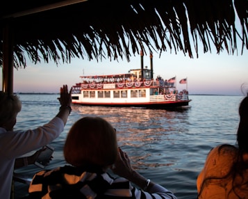 A paddle steamboat adorned with American flags and bunting sails across a calm body of water during sunset. Passengers are visible on the boat, and two people on a nearby dock wave at the passing ship, one holding a phone.