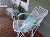 A cozy porch setting featuring a white wicker chair with a light blue and white patterned cushion. A rustic metal trunk on the left acts as a side table, holding a green vase, candle holders, a coffee mug, and a smartphone. The floor is made of wooden planks, and there's a decorative piece with a deer motif in the background.
