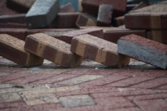 A pile of bricks is scattered on the ground, with some bricks appearing to be unevenly stacked or toppled over. The bricks have a textured surface and are mostly reddish-brown in color.
