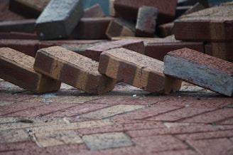 A pile of bricks is scattered on the ground, with some bricks appearing to be unevenly stacked or toppled over. The bricks have a textured surface and are mostly reddish-brown in color.