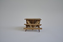 A small wooden table or bench model with a simple design, placed on a plain light gray background. The focus is clear on the structure and details of the woodwork.