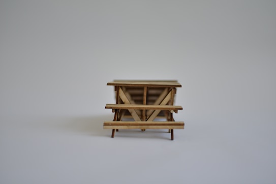 A small wooden table or bench model with a simple design, placed on a plain light gray background. The focus is clear on the structure and details of the woodwork.