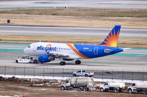 A commercial airplane with a blue and orange tail is being towed on an airport runway. The aircraft is positioned to the left with maintenance vehicles nearby. The background includes a grassy field and multiple runway markings.