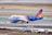 A commercial airplane with a blue and orange tail is being towed on an airport runway. The aircraft is positioned to the left with maintenance vehicles nearby. The background includes a grassy field and multiple runway markings.