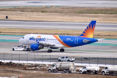 A commercial airplane with a blue and orange tail is being towed on an airport runway. The aircraft is positioned to the left with maintenance vehicles nearby. The background includes a grassy field and multiple runway markings.