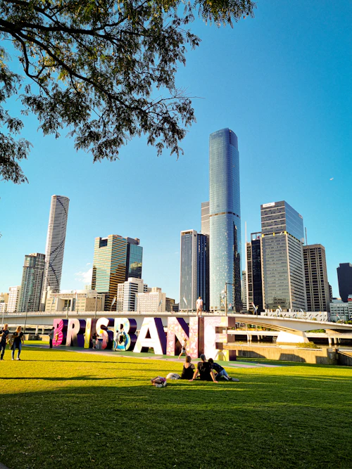 People sitting on a grassy riverside lawn with Brisbane high-rise buildings behind them