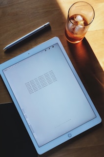 A digital tablet displaying repeated text 'Slow does not always mean slow' is placed on a wooden surface next to a stylus and a glass containing a beverage with ice cubes. The wood background is partially illuminated by sunlight.