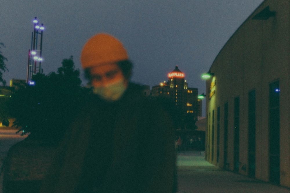 man in black jacket and orange knit cap standing on sidewalk during night time