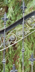 An ornate wrought iron fence featuring decorative floral patterns and fleur-de-lis accents is positioned in front of lush green fir tree branches.