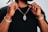 A person holding up a gold chain with a pendant, wearing a black shirt. Both wrists are adorned with bracelets, including a watch on one wrist. The focus is on the jewelry.