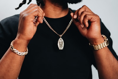 A person holding up a gold chain with a pendant, wearing a black shirt. Both wrists are adorned with bracelets, including a watch on one wrist. The focus is on the jewelry.