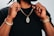 A person holding up a gold chain with a pendant, wearing a black shirt. Both wrists are adorned with bracelets, including a watch on one wrist. The focus is on the jewelry.