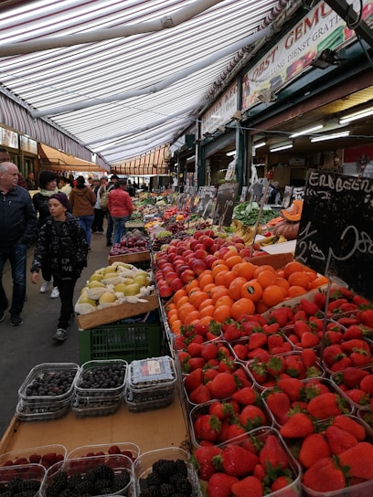 A bustling market scene with a variety of fresh fruits on display. The stall is arranged with vibrant fruits such as strawberries, oranges, and pears, neatly placed in rows. Shoppers are seen walking under a striped canopy roof. Signage with pricing is visible, adding to the lively market atmosphere.